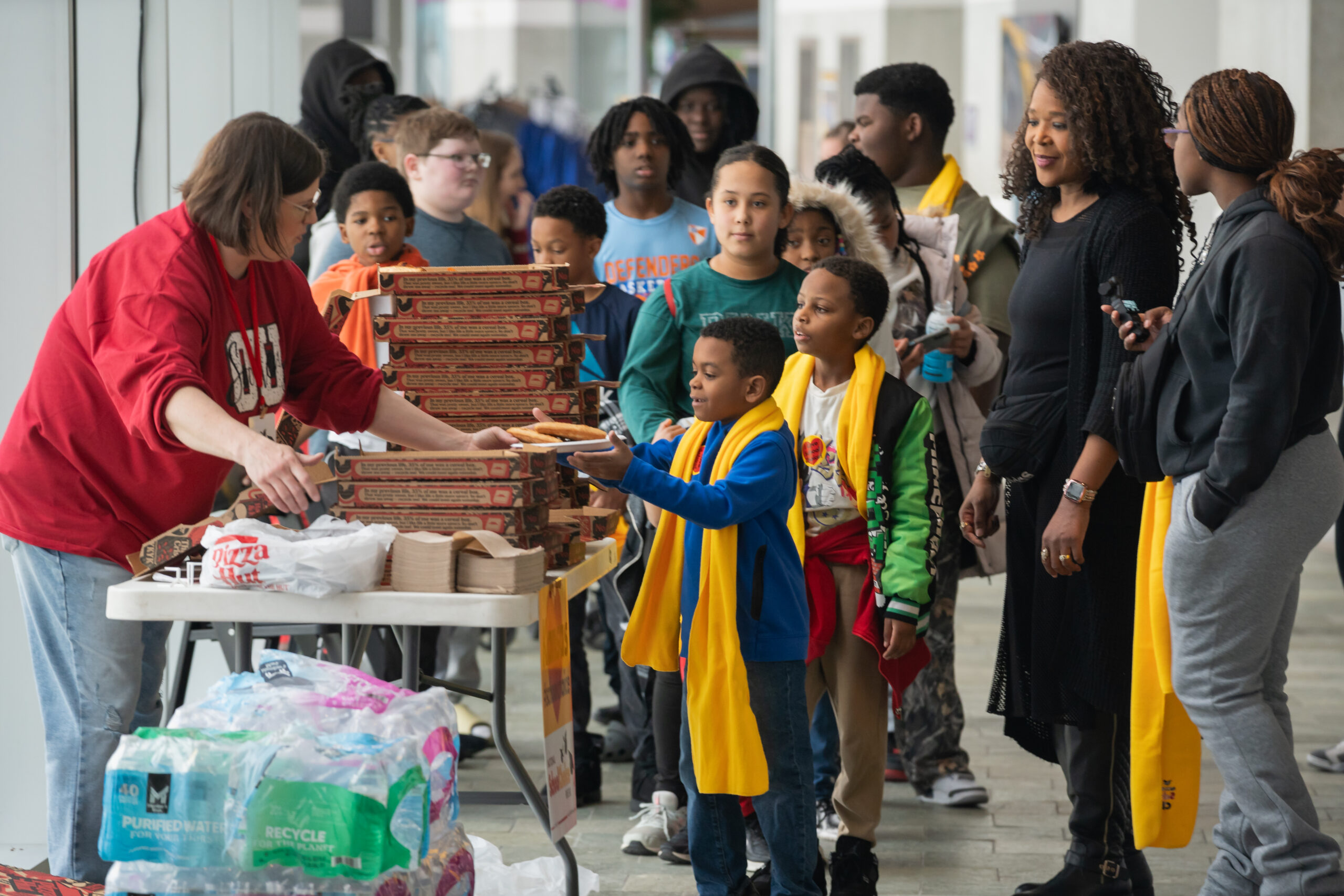 A grade-school student wearing a scarf grabs a plate of pizza at an event